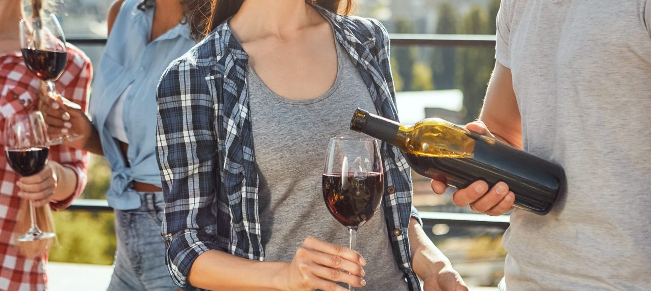 Having fun with friends. Young bearded men is pouring wine to his friend and smiling while standing on the roof with friends.