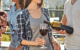 Having fun with friends. Young bearded men is pouring wine to his friend and smiling while standing on the roof with friends.