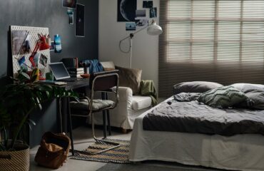 Double bed with grey cushions and blanket standing in front of desk with computer, photos and books in spacious bedroom of teenager